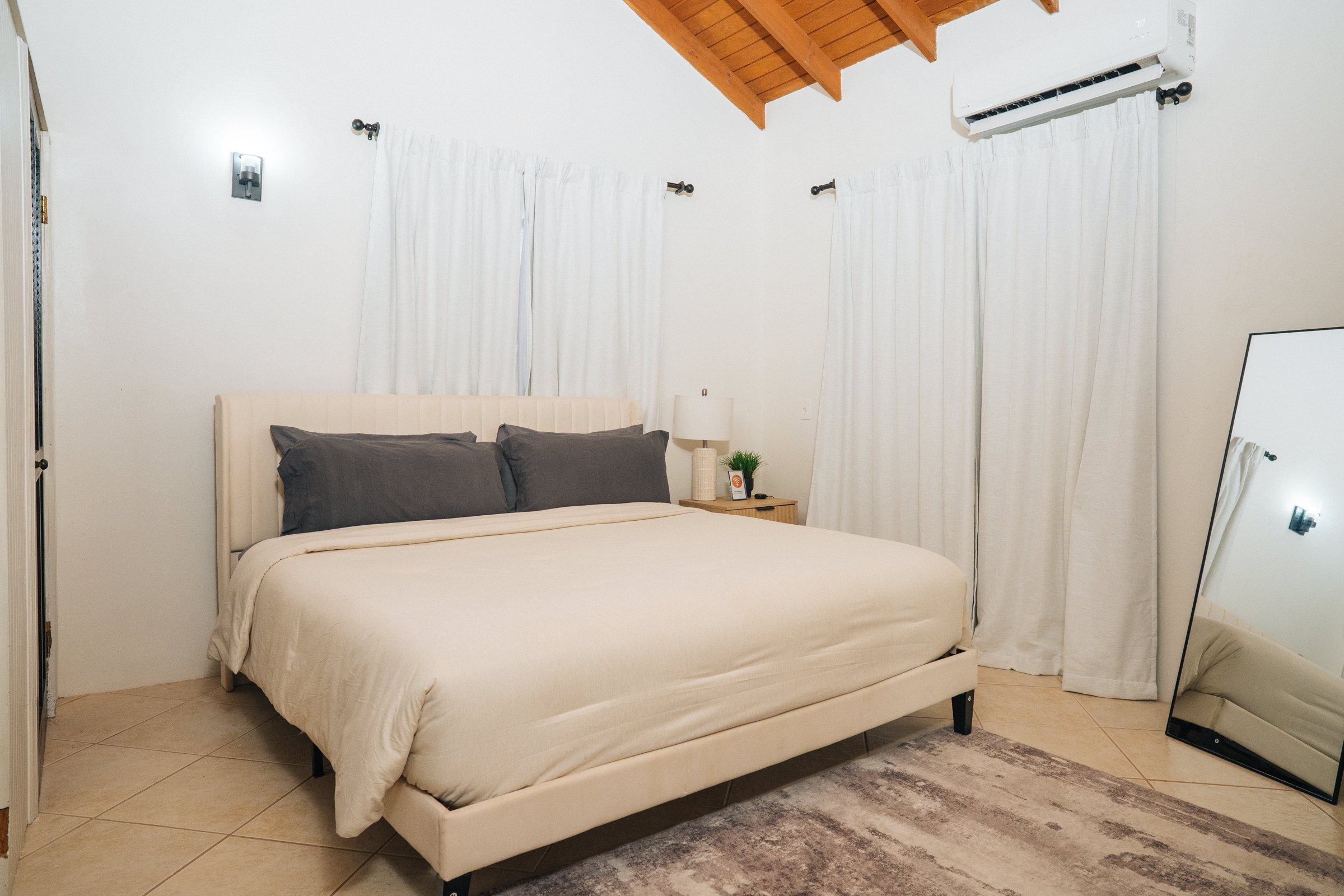 Cream upholstered bed with soft bedding, wooden ceiling beams, and air conditioning